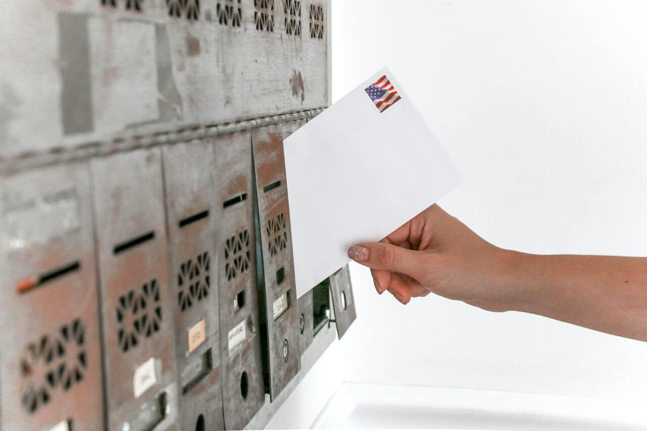 Hand slipping a white envelope into a gray metal mailbox