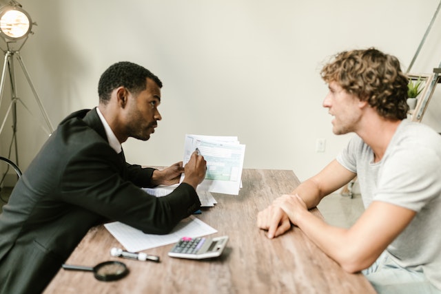 A person in a suit holding a printed paper and pointing something out to the person sitting across from them